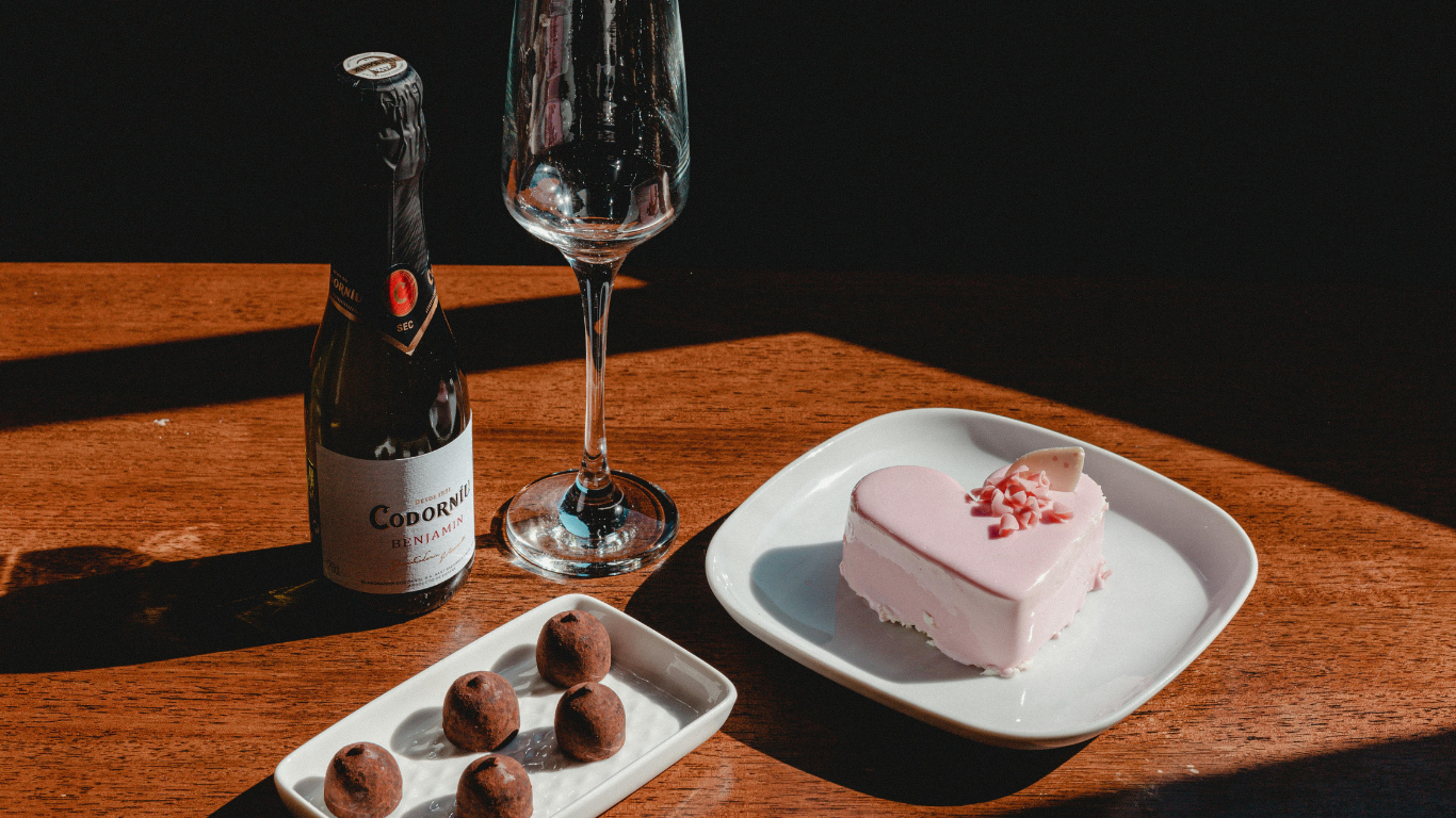 Small bottle of sparkling wine, an empty champagne flute, chocolate truffles and a pink heart‑shaped dessert arranged on a wooden table in warm light.