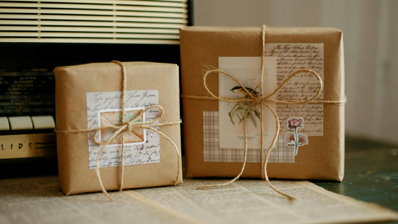 Two gifts wrapped in brown kraft paper and tied with twine, decorated with vintage‑style paper, handwriting, and floral stickers, sitting on an open book or newspaper in a cozy, nostalgic setting.