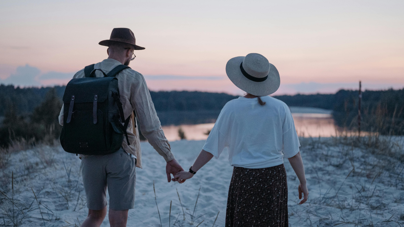 Couple walking hand in hand over sandy ground toward a lake at sunset, both wearing hats and casual clothes, with a backpack on one person, suggesting a relaxed romantic getaway.