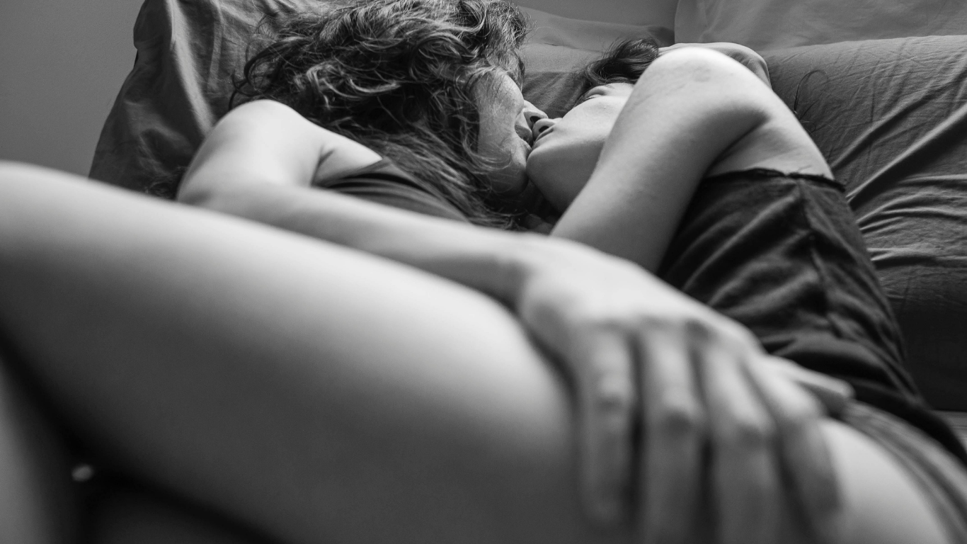 Couple lying in bed facing each other, wrapped in white sheets with soft warm lighting, touching gently and smiling while sharing an intimate, romantic moment.