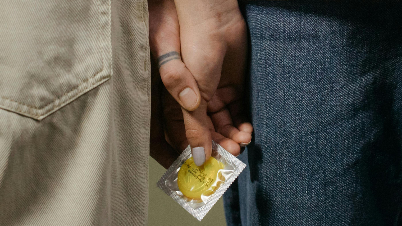 Close‑up of two people standing side by side, holding hands, with one person discreetly holding a yellow condom packet between their fingers, symbolising first time sex, safety and consent.