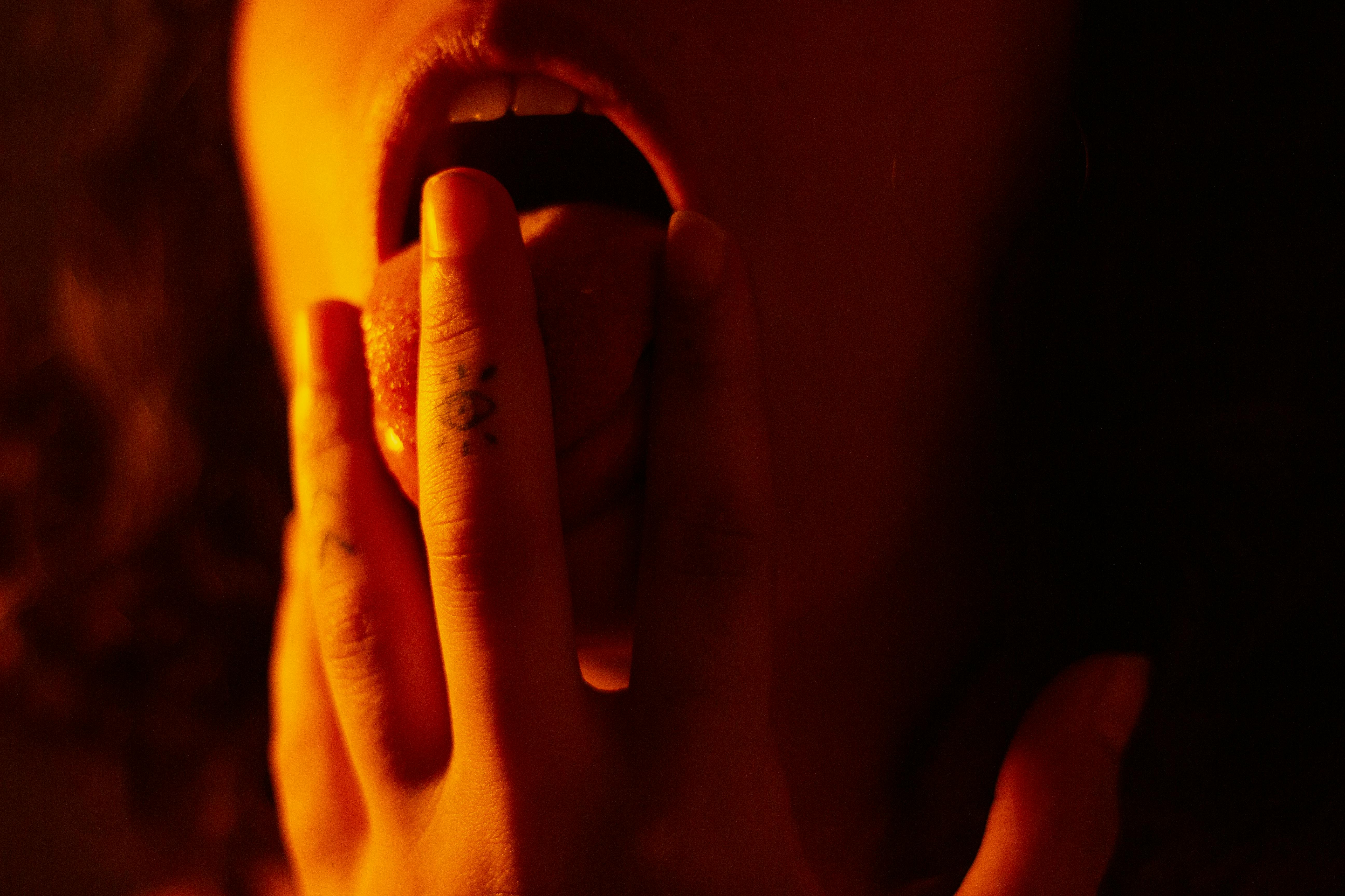 Close-up of an open mouth with tattooed fingers touching the lips, bathed in warm amber light, evoking sensual touch and oral intimacy