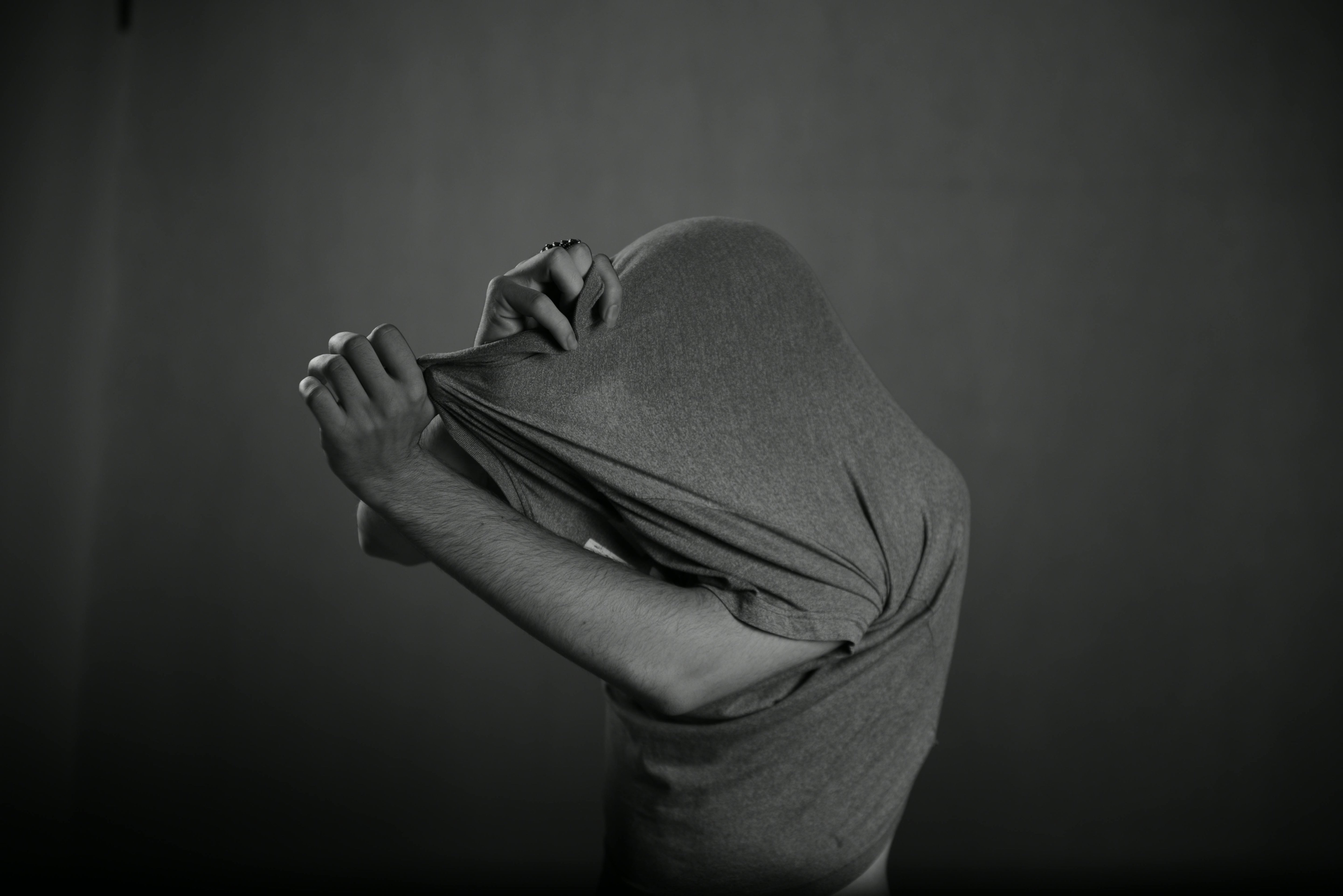 Black and white photo of a person pulling a T‑shirt over their head, with their face completely covered and only their bare arms visible against a plain dark background.